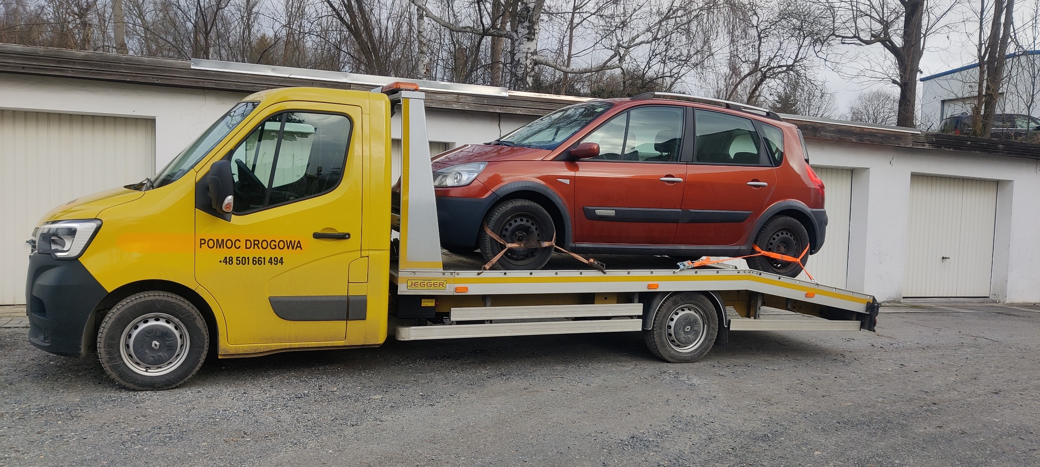 Żółta laweta z pomarańczowym samochodem osobowym zabezpieczonym pasami transportowymi, na tle garaży.