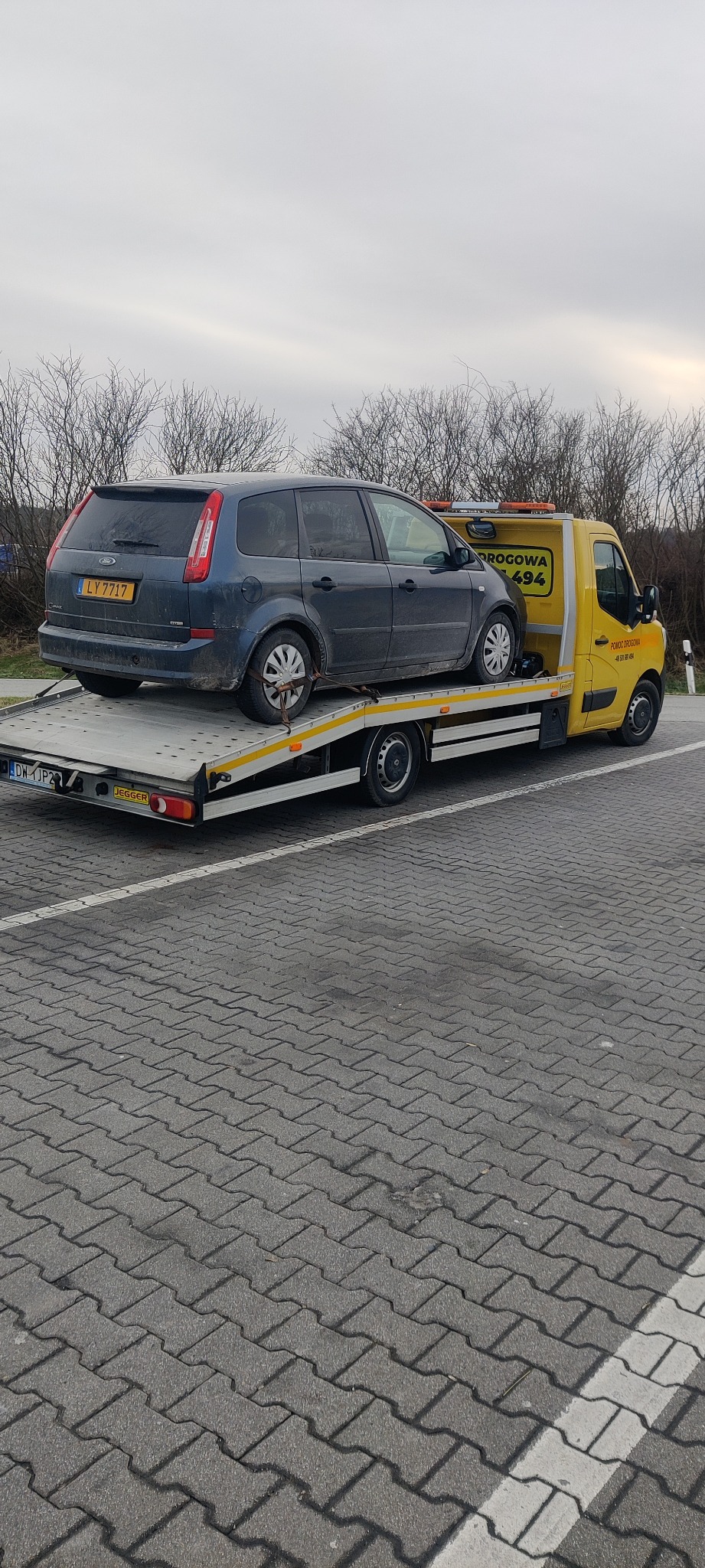 Niebieski samochód osobowy typu kombi, zabezpieczony pasami, transportowany na żółtej lawecie pomocy drogowej na brukowanym parkingu.