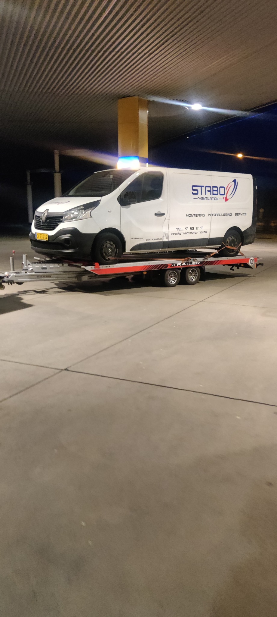 Biały samochód dostawczy z logo firmy STABO Ventilation przewożony na czerwonej lawecie pod zadaszeniem stacji benzynowej, widoczne niebieskie światła ostrzegawcze na dachu.