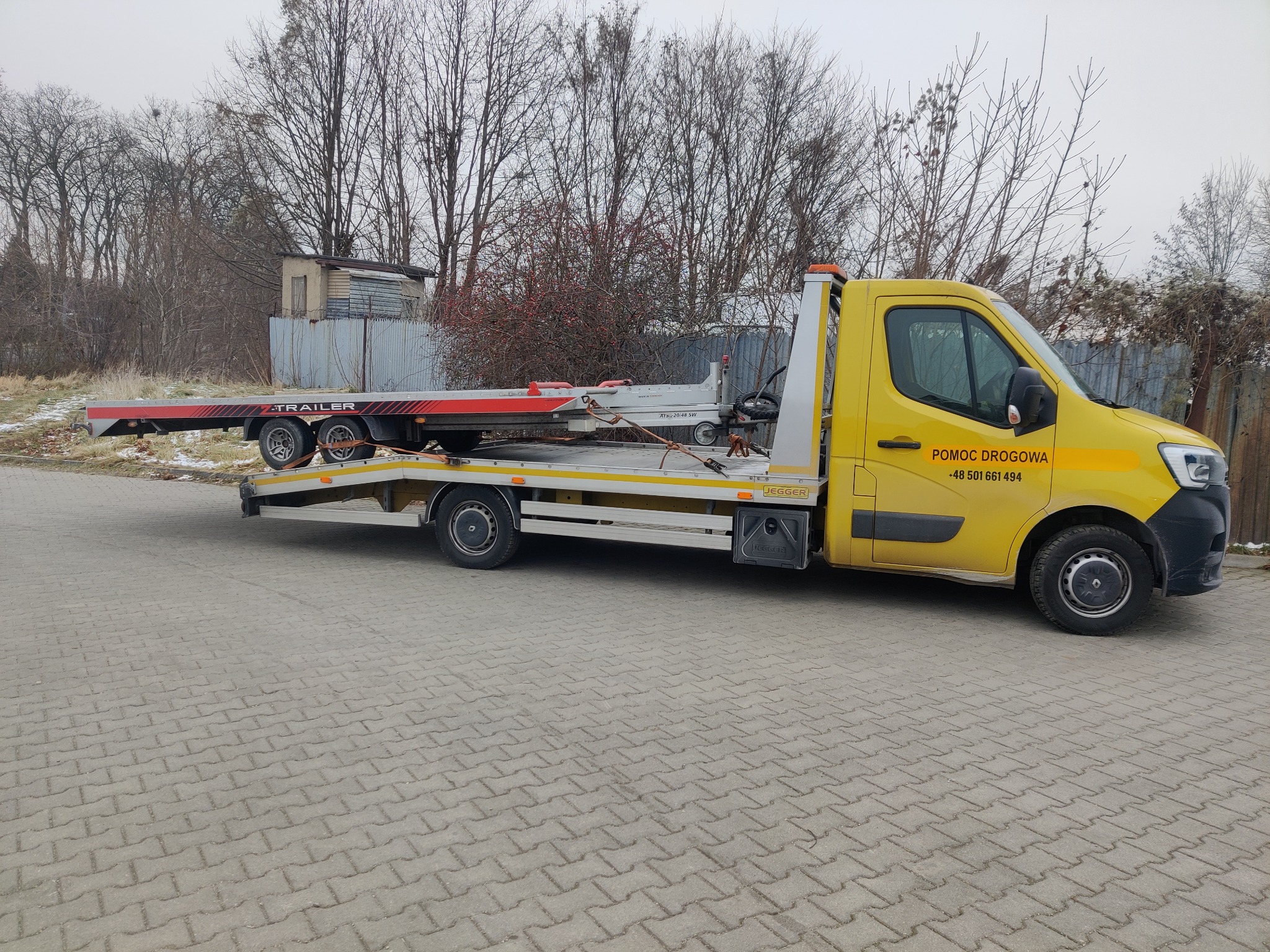 Żółta laweta z przyczepą na tle drzew i szarej kostki brukowej. Na boku pojazdu napis 'Pomoc Drogowa' i numer telefonu.