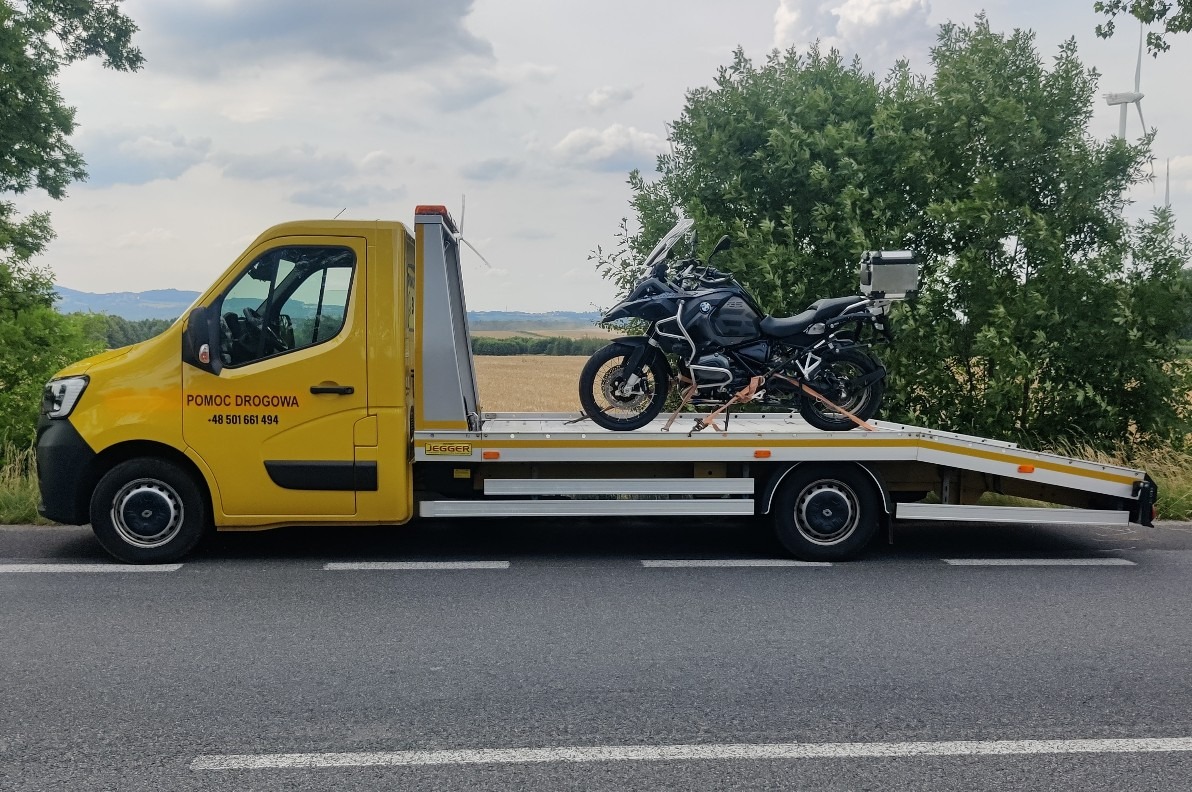 Żółta laweta z motocyklem BMW zabezpieczonym pasami transportowymi, zaparkowana na poboczu drogi, w tle krajobraz z turbinami wiatrowymi.