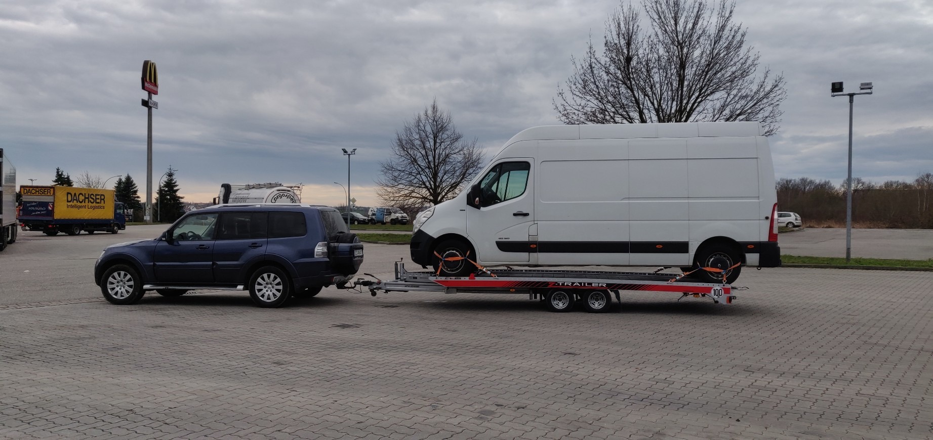 Niebieski SUV ciągnie lawetę z białym busem w tle parkingu z widocznym znakiem McDonald's.