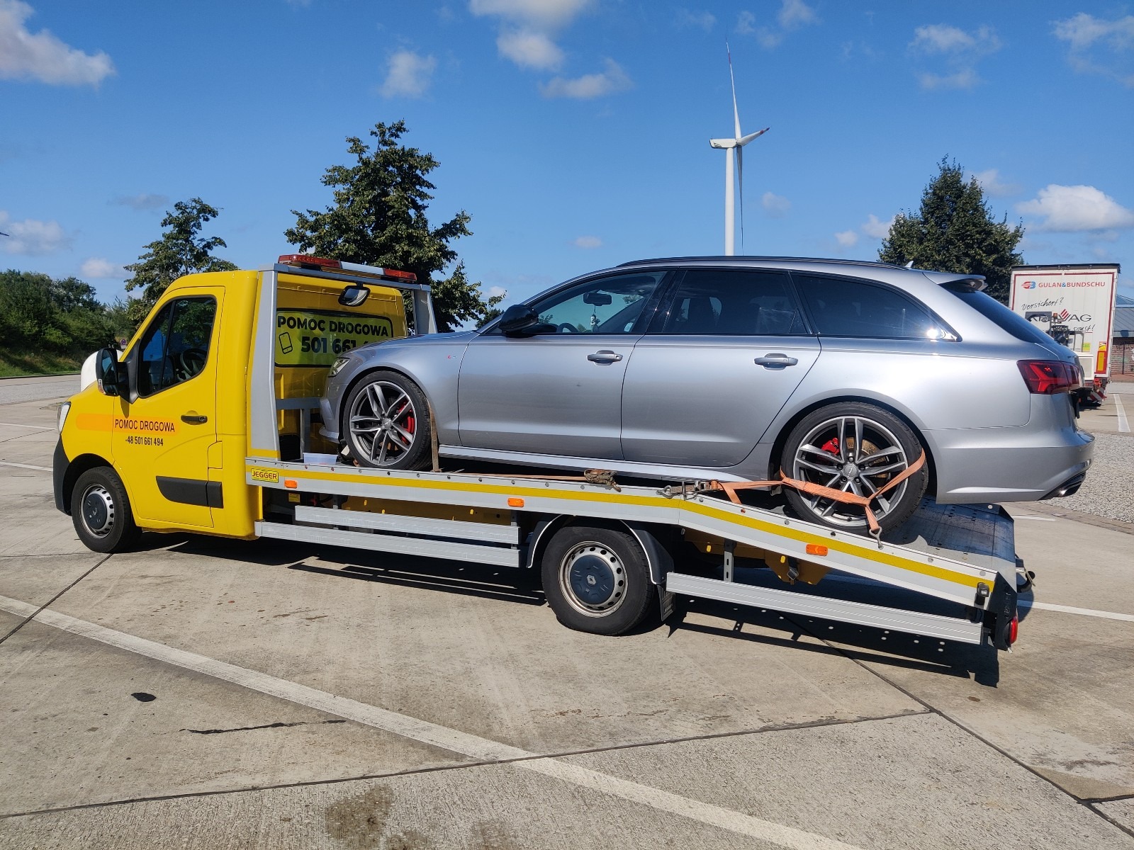 Żółta laweta z srebrnym Audi kombi na platformie, zabezpieczonym pomarańczowymi pasami, na parkingu w słoneczny dzień.