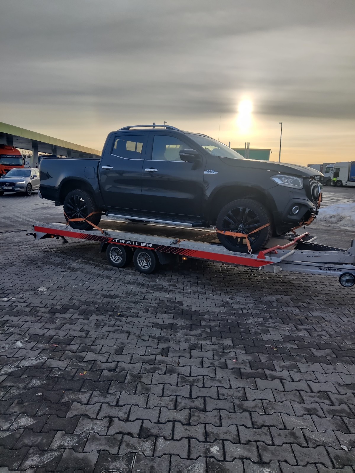 Czarny pickup Mercedes-Benz X-Class zabezpieczony pomarańczowymi pasami na czerwono-szarej lawecie dwuosiowej na brukowanym placu, w tle budynek stacji benzynowej i inne pojazdy.