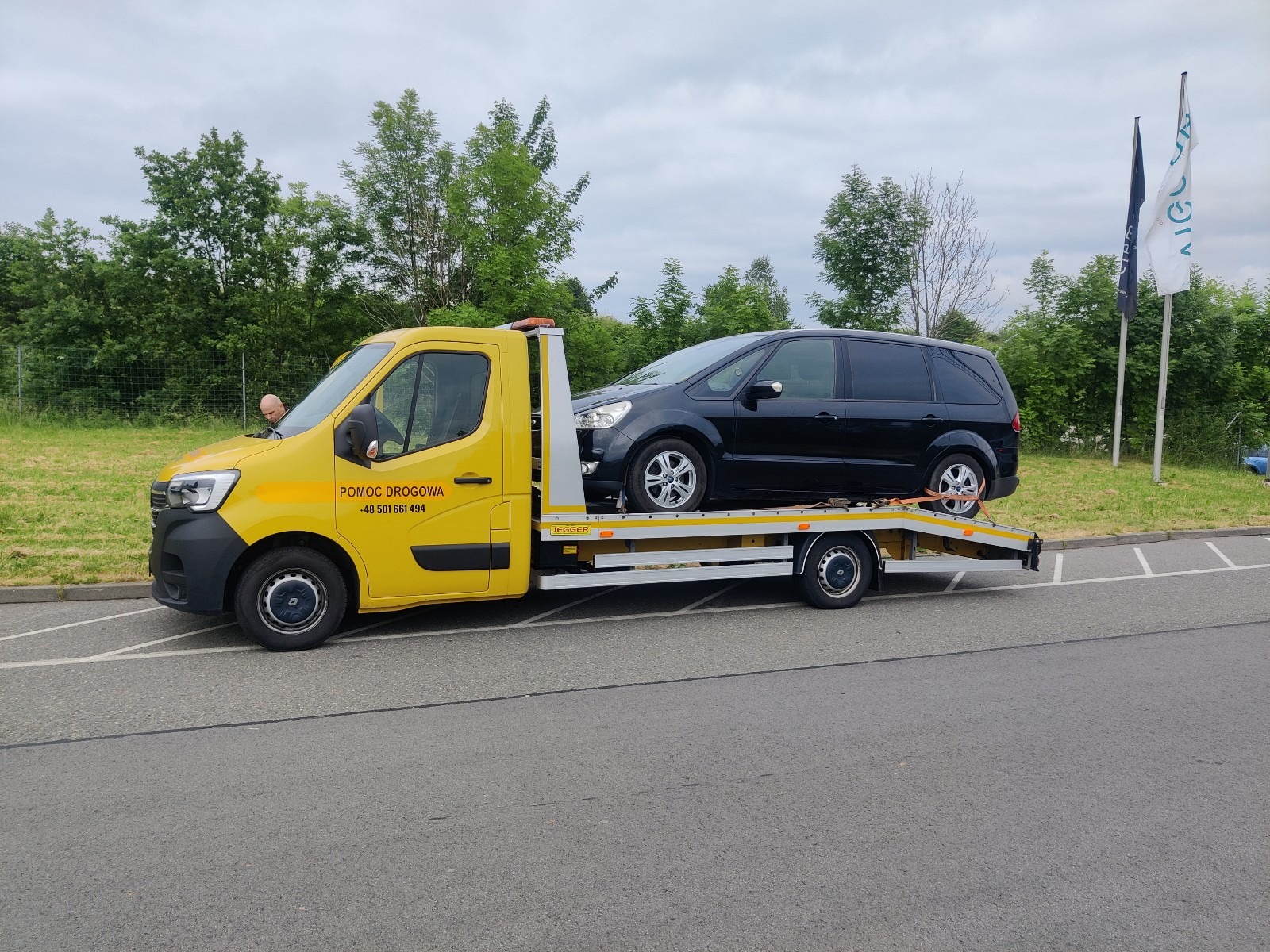 Żółta laweta z czarnym samochodem osobowym na platformie, zabezpieczonym pasami transportowymi, widoczny kierowca lawety