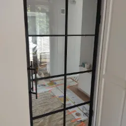 Czarna, metalowa rama drzwi szklanych z podziałami w stylu loft, widok na zabezpieczoną podłogę i wnętrze pomieszczenia w trakcie remontu.