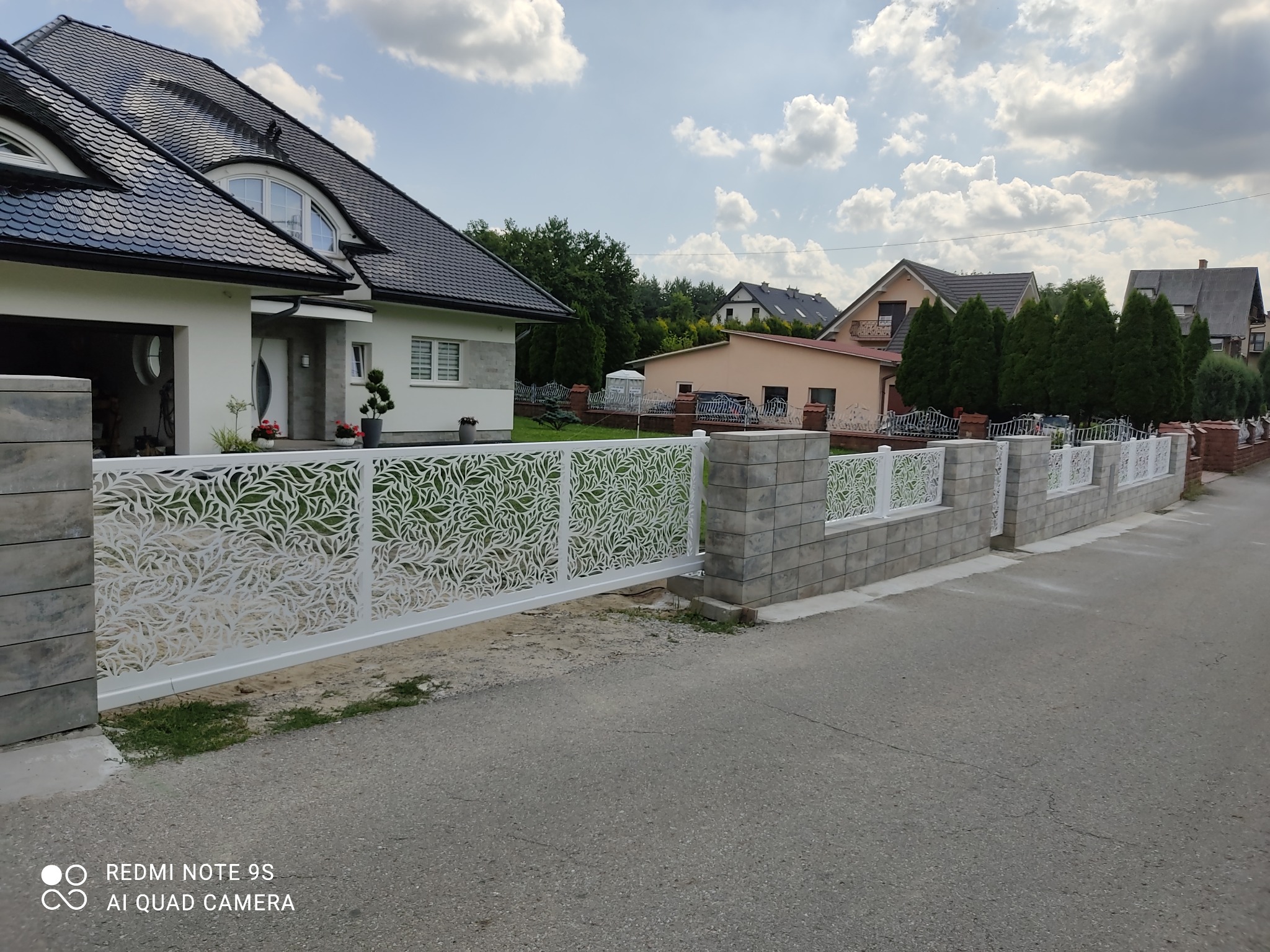 Nowoczesna, biała brama wjazdowa z laserowo wycinanym wzorem liści, osadzona na szarych, betonowych słupkach, na tle domów jednorodzinnych i błękitnego nieba.