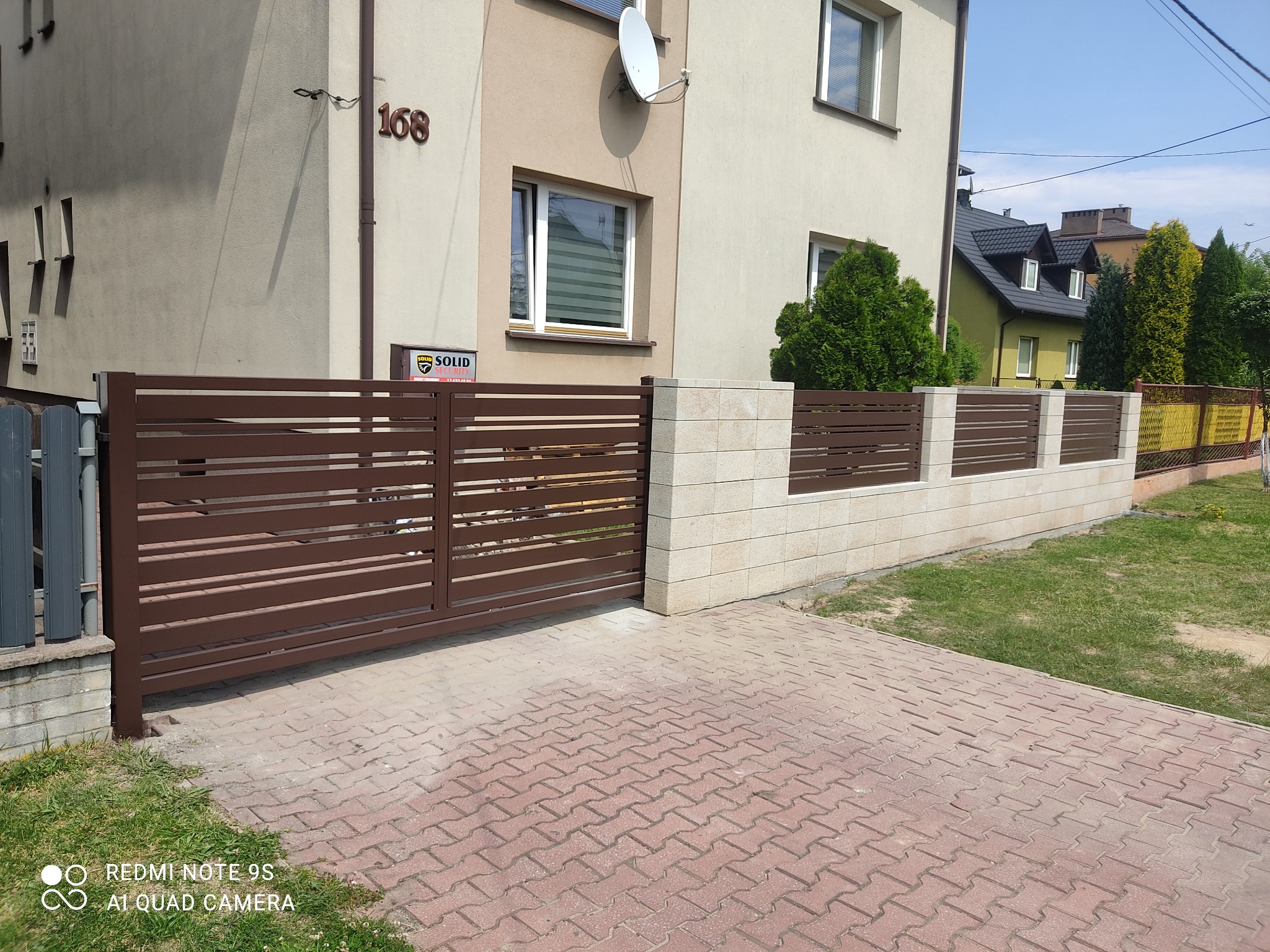 Automatyczna brama przesuwna z poziomymi profilami w kolorze brązowym, zamontowana przy posesji z numerem 168, widoczna kostka brukowa i fragment trawnika.