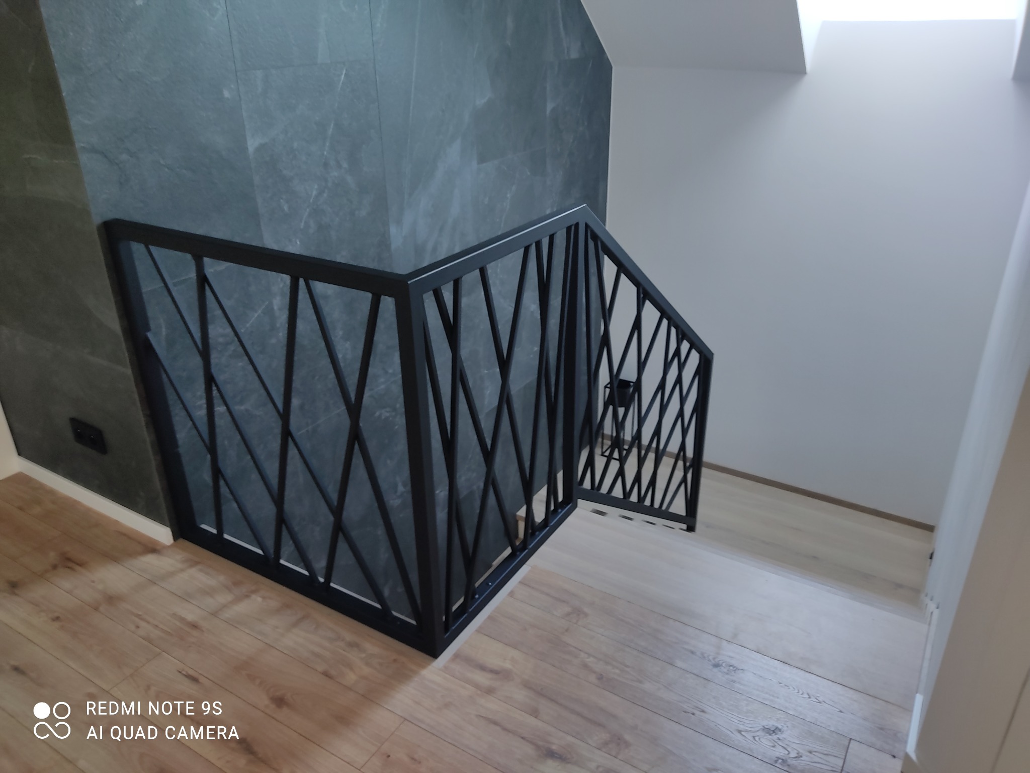 Nowoczesne schody wewnętrzne z czarną, metalową balustradą o geometrycznym wzorze, widoczne na tle szarej ściany z efektem kamienia i drewnianej podłogi.