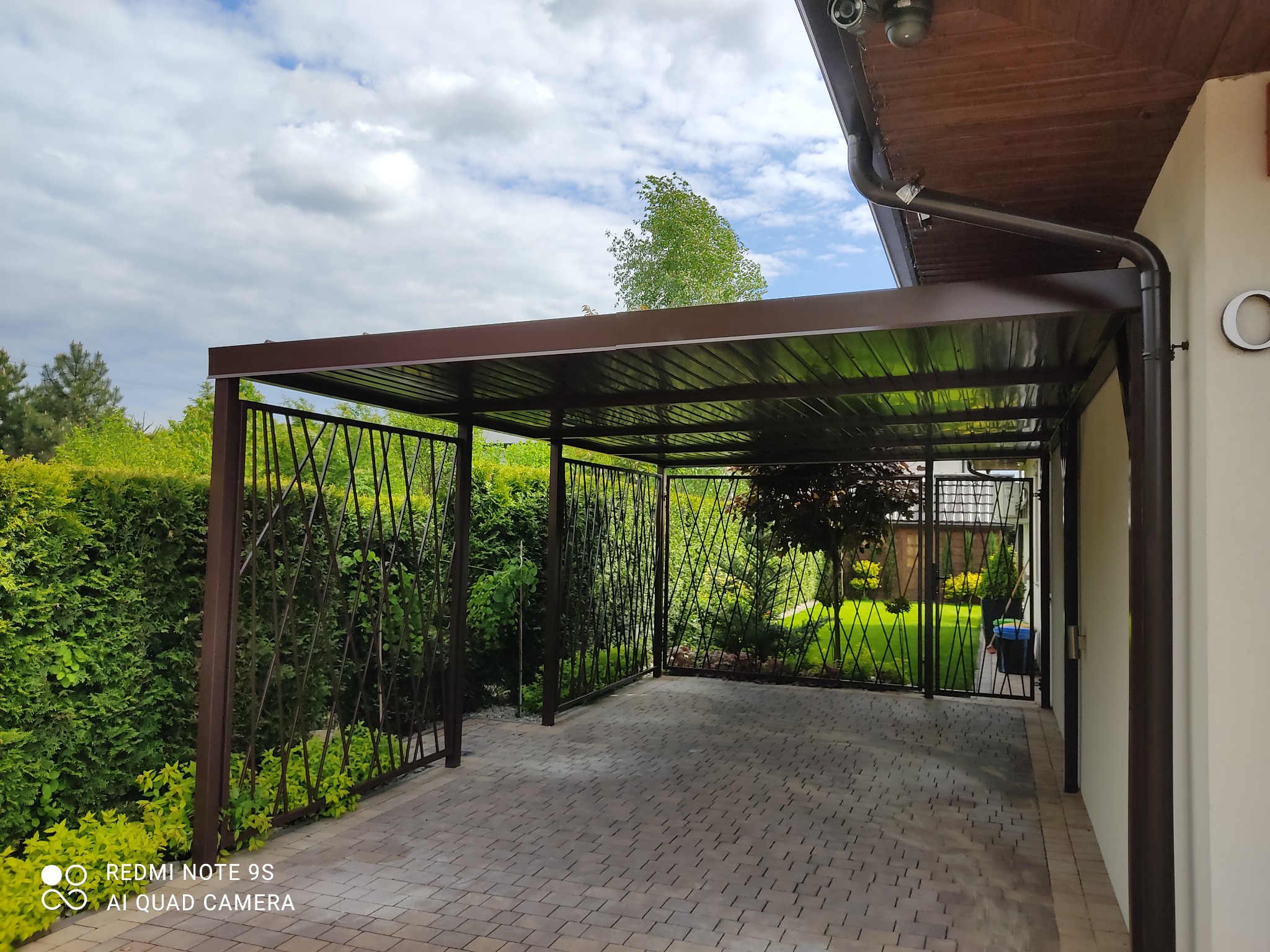 Zadaszone miejsce parkingowe z ażurowymi ścianami, brązowa konstrukcja na kostce brukowej, widok na zadbany ogród i fragment budynku.