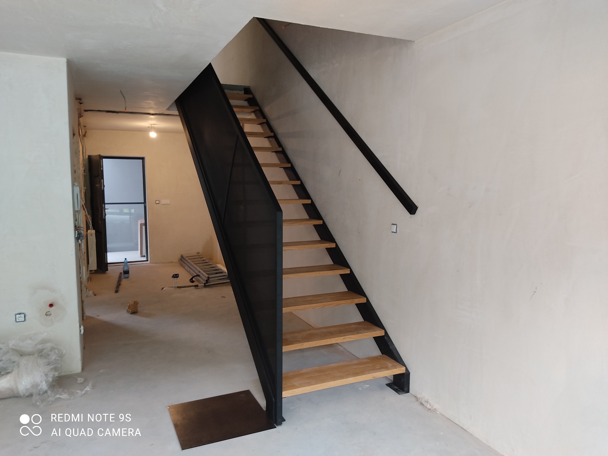 Metalowe schody z drewnianymi stopniami i czarną balustradą w surowym wnętrzu, widoczne ślady prac wykończeniowych na ścianach i podłodze.
