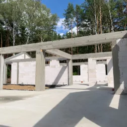 Surowy szkielet powstającego budynku z betonu komórkowego, z widocznymi betonowymi nadprożami i zbrojeniami, na tle lasu i błękitnego nieba.