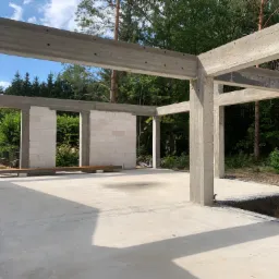 Surowy stan domu w Wyszkowie: widok na betonowy fundament, ściany z bloczków i konstrukcję szkieletową, otoczone zielenią drzew.