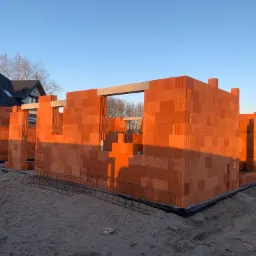 Wznoszone ściany z czerwonej ceramiki poryzowanej w stanie surowym otwartym, widoczne zbrojenia fundamentów, w tle dom jednorodzinny.