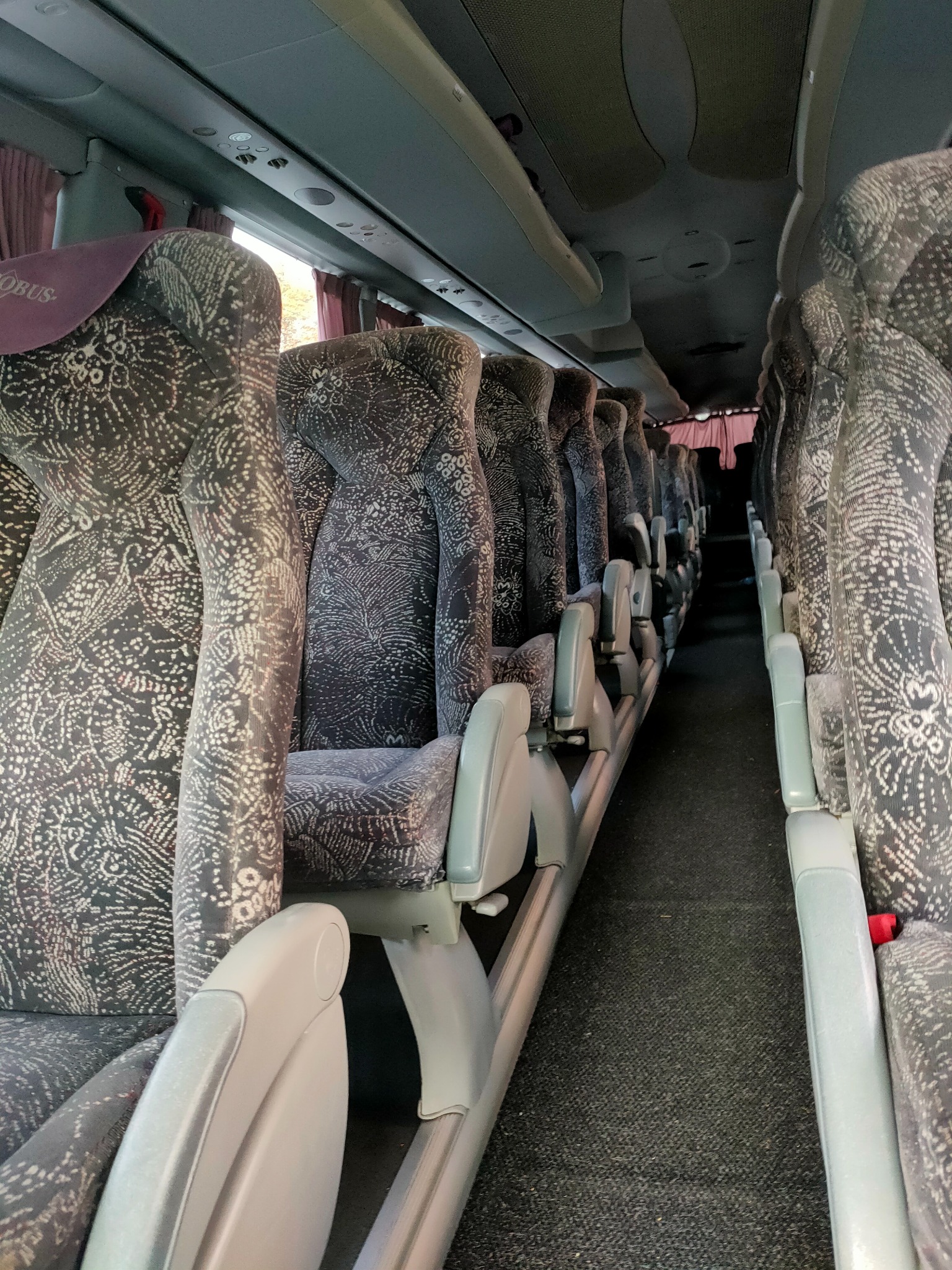 Wnętrze autobusu turystycznego z rzędami foteli obitych ciemną tkaniną w jasny wzór, widok z perspektywy pasażera.