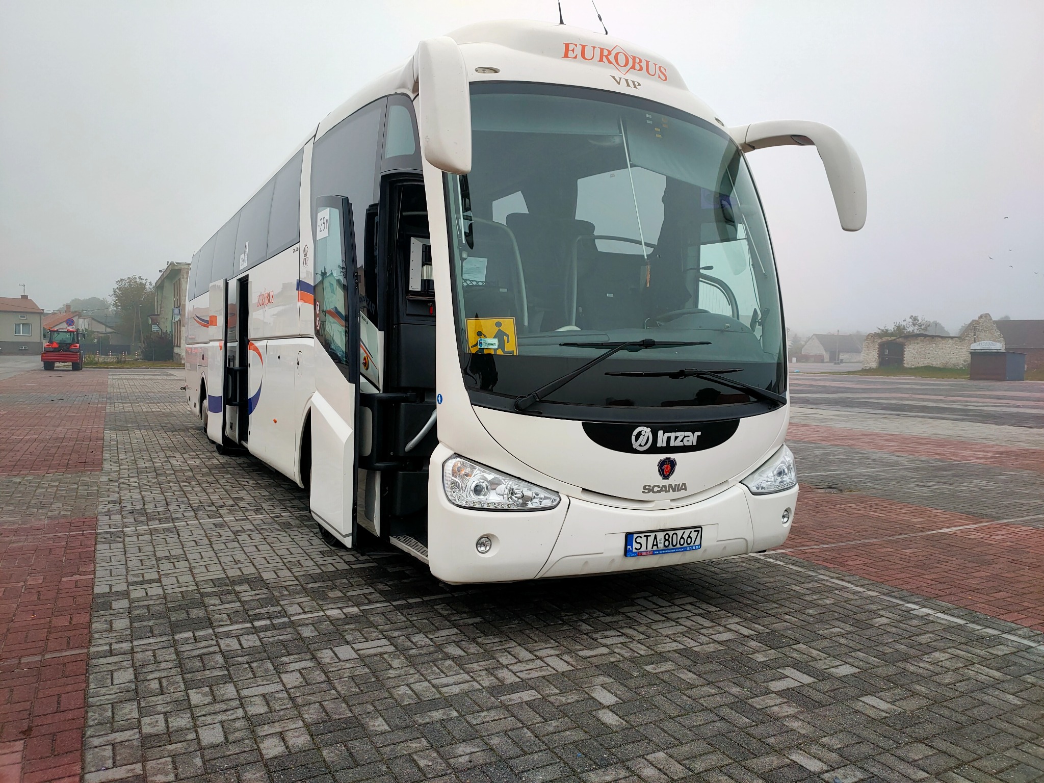 Biały autokar Eurobus VIP marki Irizar Scania z otwartymi drzwiami, zaparkowany na placu z kostki brukowej w pochmurny dzień.