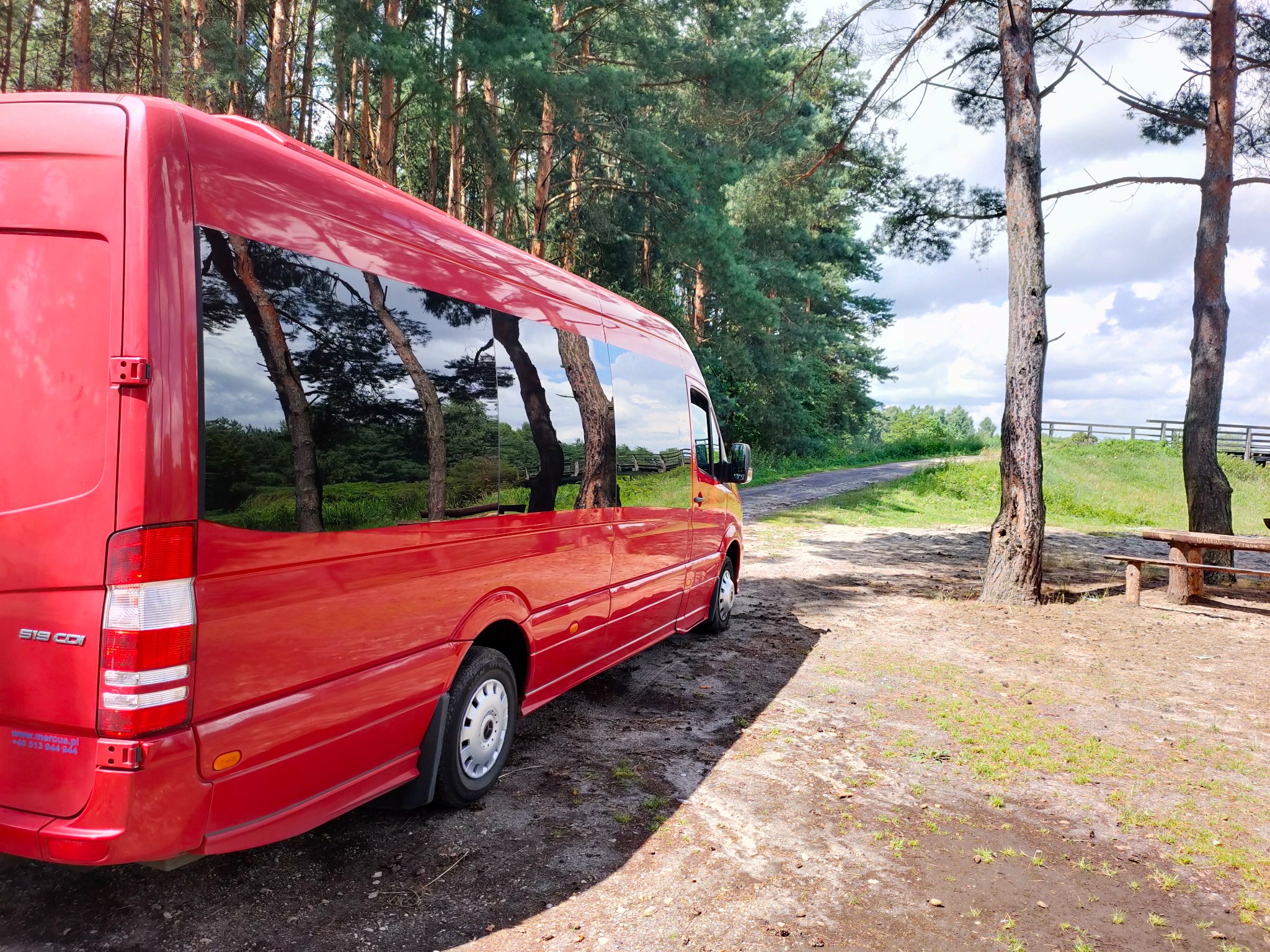 Czerwony bus marki Mercedes-Benz Sprinter z przyciemnianymi szybami, zaparkowany na leśnej drodze obok drewnianej ławki.