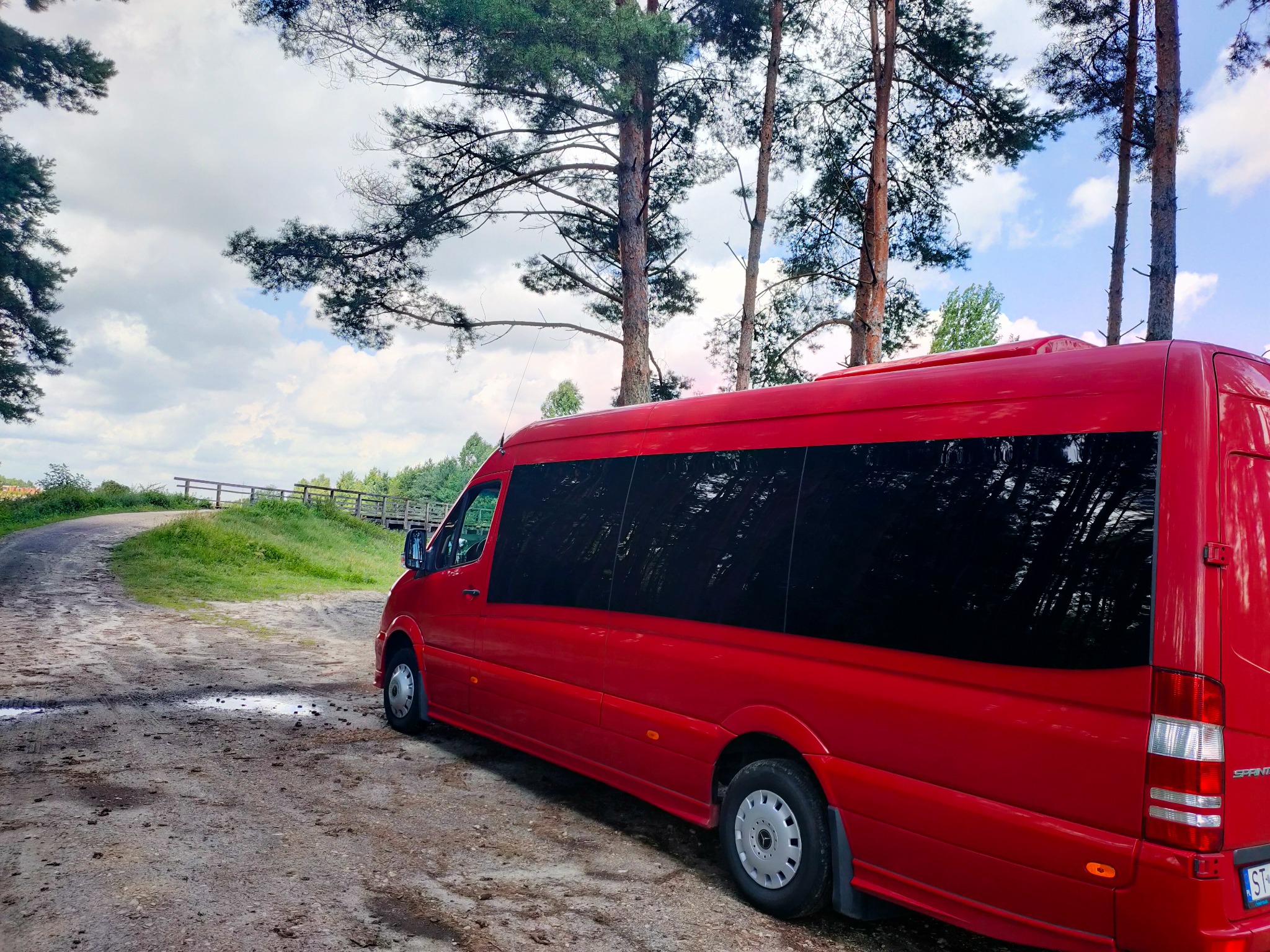 Czerwony bus z przyciemnianymi szybami zaparkowany na leśnej drodze, w tle widoczne drzewa i fragment drewnianego mostku.