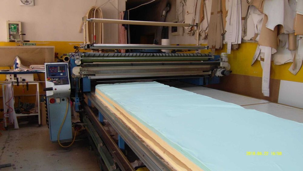 Automatyczna krajarka tnąca warstwy jasnoniebieskiej tkaniny w zakładzie odzieżowym, z papierowymi wykrojami wiszącymi na ścianie w tle.