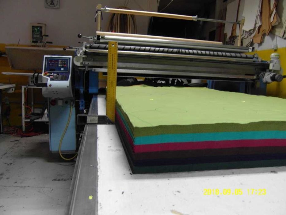 Wielowarstwowa sterta kolorowych tkanin pod automatyczną maszyną do cięcia, z żółtą miarką pionowo przy krawędzi, widoczne zarysy wzorów do wycięcia na wierzchniej warstwie.