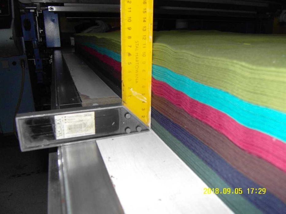 Wielobarwny stos tkanin układany i wyrównywany na stole krojczym z użyciem kątownika stalowego z podziałką centymetrową.