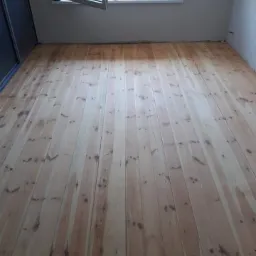 Surowa podłoga z jasnego drewna sosnowego, świeżo ułożona w pustym pokoju z otwartym oknem i widokiem na zieleń, przed wykończeniem.