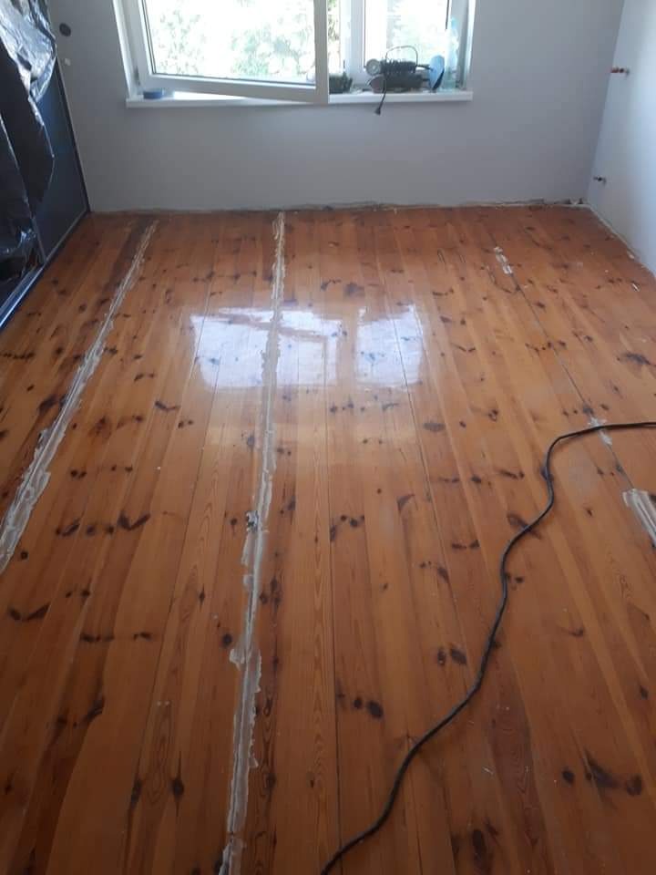 Lakierowana podłoga z desek, częściowo wypełniona szpachlą, widoczny kabel elektryczny i okno z narzędziami na parapecie.