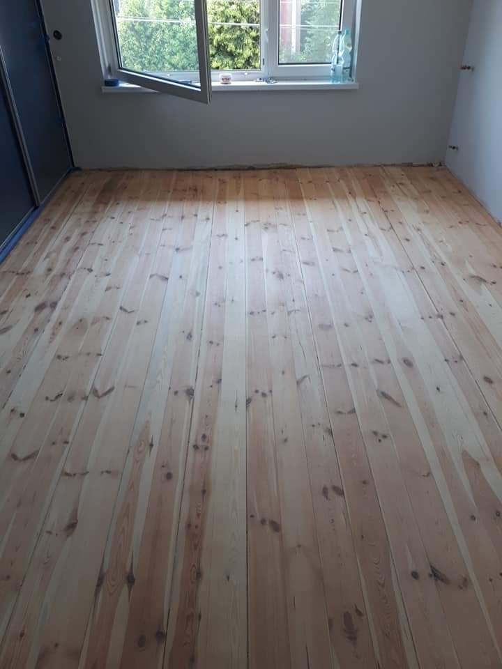 Surowa podłoga z jasnego drewna sosnowego, świeżo ułożona w pustym pokoju z otwartym oknem i widokiem na zieleń, przed wykończeniem.