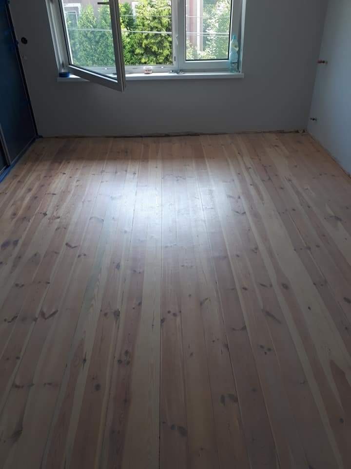 Świeżo ułożona podłoga z jasnych paneli drewnianych w pustym pokoju z otwartym oknem, widoczne ślady po taśmie malarskiej na ścianach.