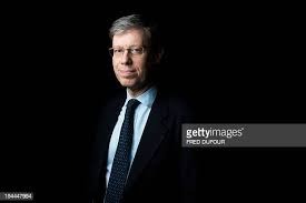 Portret mężczyzny w okularach i ciemnym garniturze z krawatem w kropki na czarnym tle.