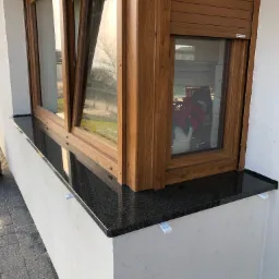 Czarny, błyszczący element wykończeniowy pod oknem z drewnianą ramą i roletą zewnętrzną, zamontowany na elewacji budynku, widoczne białe wsporniki montażowe.