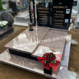 Kamieniarstwo Robert Didyk - Wielopoziomowy pomnik nagrobny z brązowego kamienia z czarnymi tablicami inskrypcyjnymi, dekorowany bukietem czerwonych róż i metalowym krzyżem z figurą Chrystusa, na tle cmentarza.