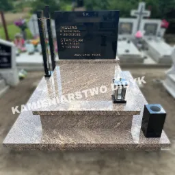 Granitowy pomnik nagrobny z czarną tablicą inskrypcyjną, metalowym krzyżem i lampionem, na tle innych nagrobków i zieleni cmentarza.