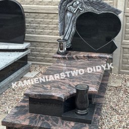 Kamieniarstwo Robert Didyk - Granitowy nagrobek z rzeźbą anioła i sercem, z ciemnego kamienia z wzorem, na żwirowym podłożu. Widoczny lampion i wazon. Tło stanowi betonowy mur.