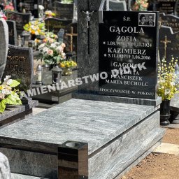 Kamieniarstwo Robert Didyk - Granitowy nagrobek z czarną tablicą inskrypcyjną i zdjęciem zmarłych, otoczony innymi nagrobkami na cmentarzu w słoneczny dzień. Widoczny krzyż i kwiaty.