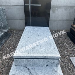 Kamieniarstwo Robert Didyk - Nowoczesny nagrobek z jasnego granitu z czarnym krzyżem na tle betonowego muru. Widoczne drobne kamienie wokół.
