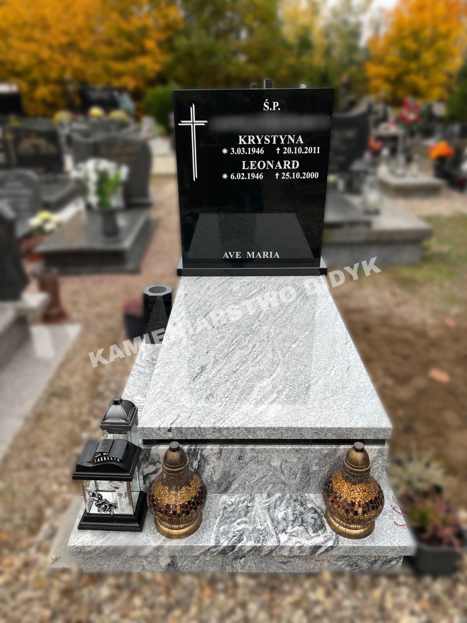 Czarny nagrobek z jasnego, wzorzystego kamienia w Bukowcu, z wygrawerowanym krzyżem i inskrypcją, otoczony lampionami i zniczami na tle cmentarza z jesiennymi drzewami.