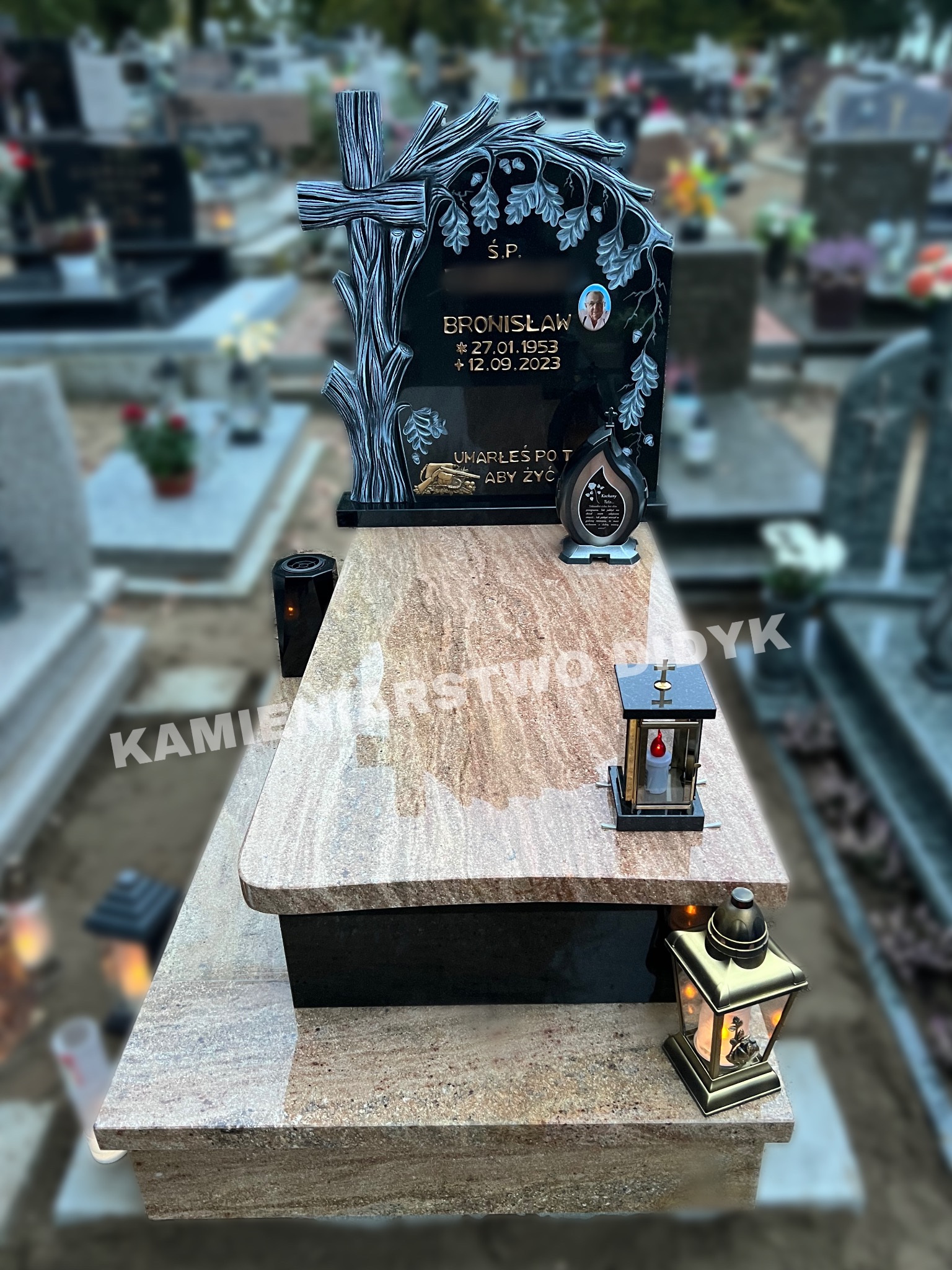 Granitowy pomnik z czarną tablicą, ozdobiony motywem drzewa i krzyża, z inskrypcją i zdjęciem zmarłego, oświetlony lampionami na cmentarzu.