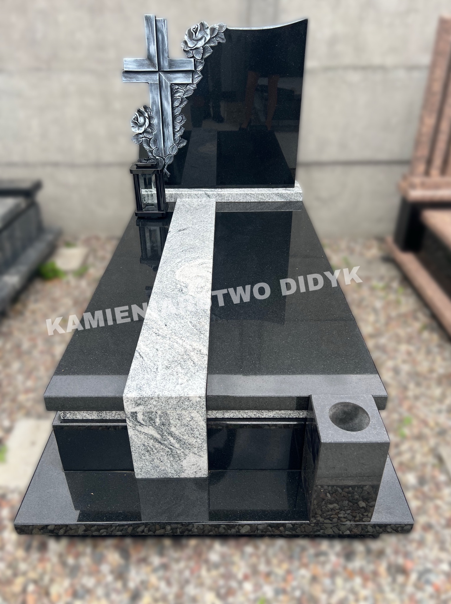Nowoczesny, czarny pomnik z szarym pasem i krzyżem z motywem róż, z lampionem, na tle szarej ściany i żwirowego podłoża.