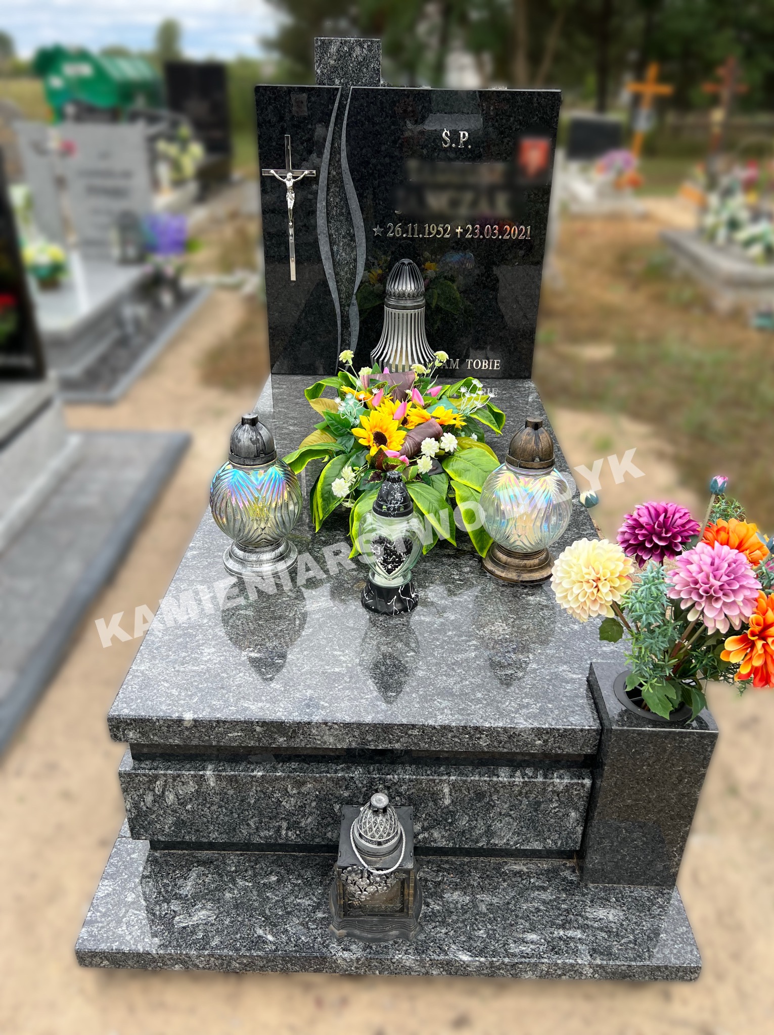 Granitowy pomnik nagrobny z krzyżem, datami i kwiatami, otoczony lampionami, na cmentarzu.