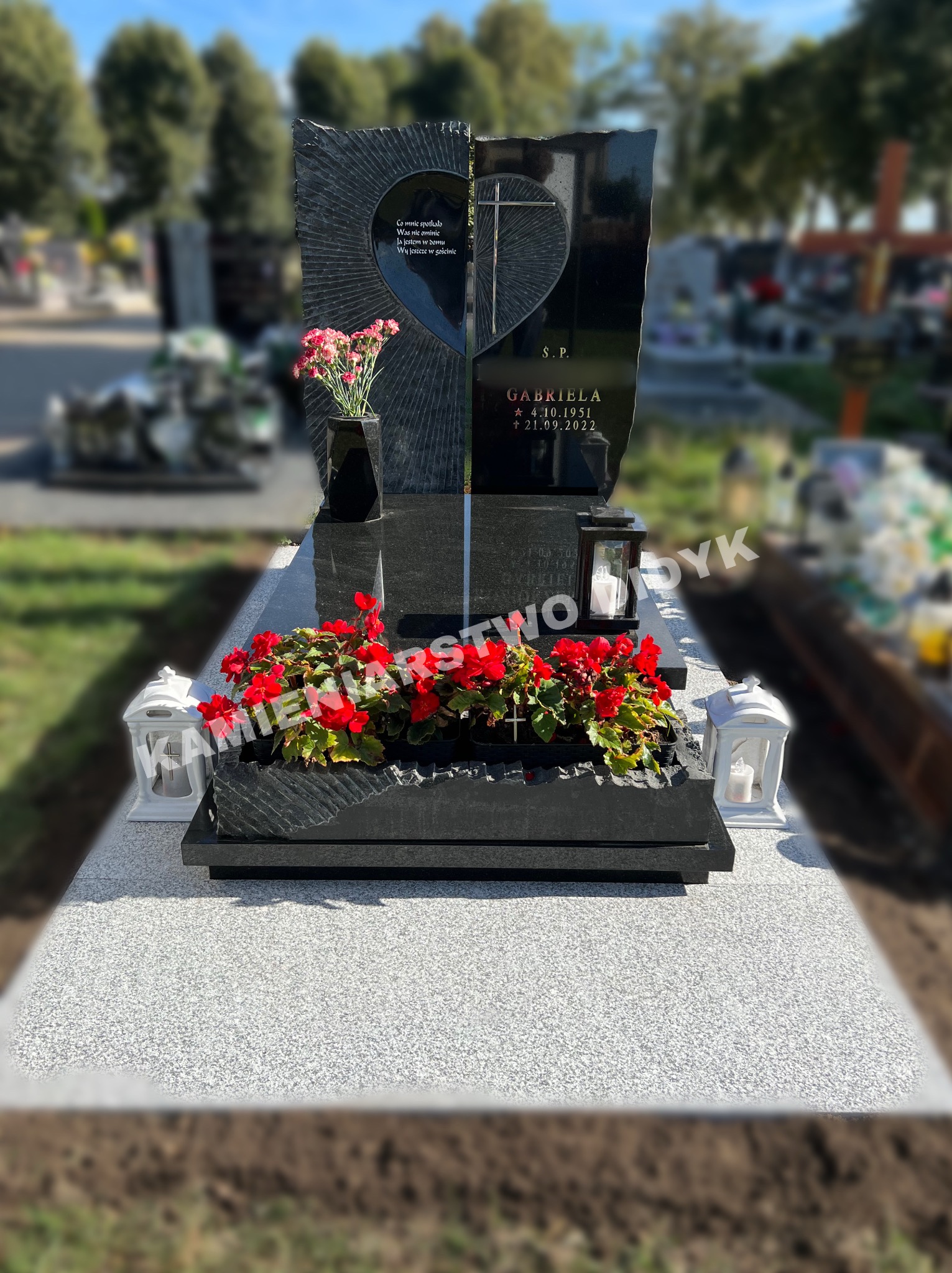 Czarny, nowoczesny pomnik z wyciętym sercem i napisem, ozdobiony świeżymi kwiatami w wazonie i czerwonymi begoniami w donicach, otoczony lampionami na cmentarzu.