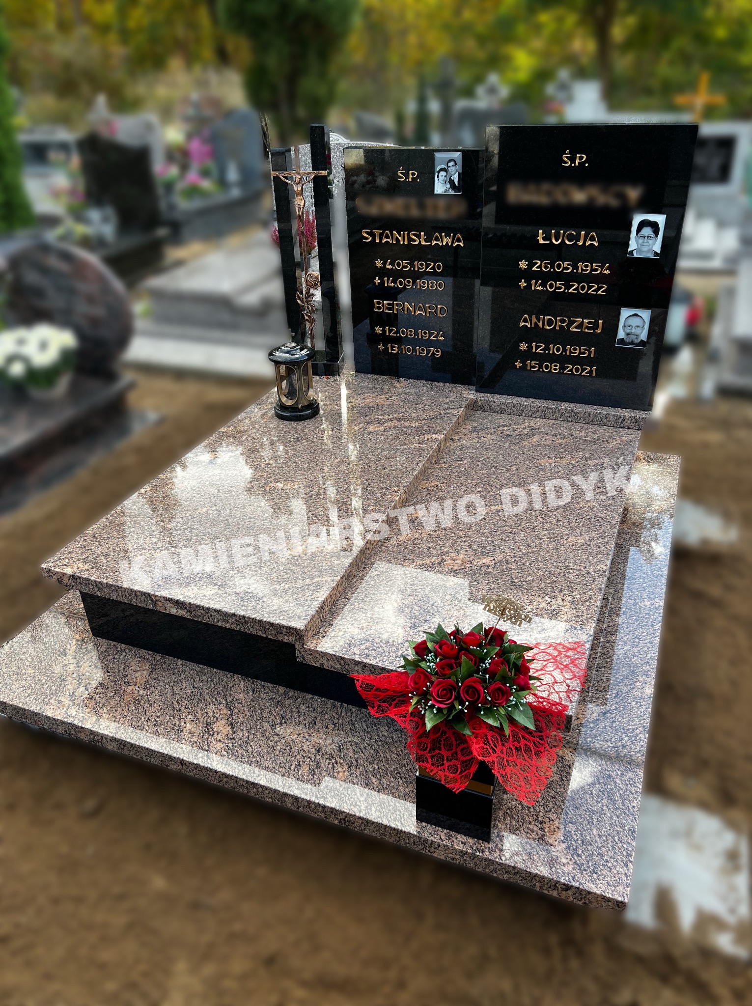 Wielopoziomowy pomnik nagrobny z brązowego kamienia z czarnymi tablicami inskrypcyjnymi, dekorowany bukietem czerwonych róż i metalowym krzyżem z figurą Chrystusa, na tle cmentarza.