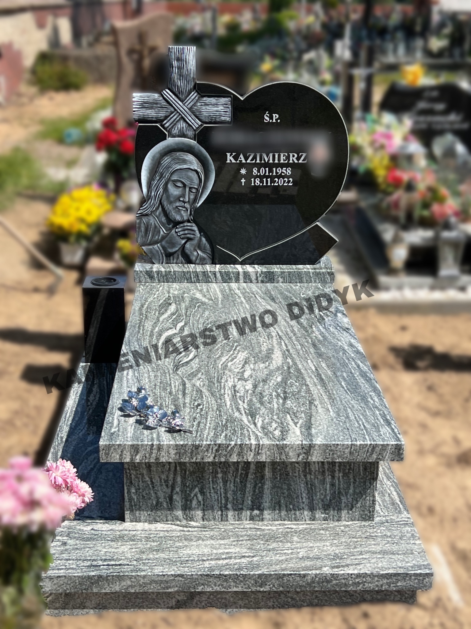 Granitowy nagrobek z czarnym sercem i wizerunkiem Jezusa, na tle innych nagrobków i zieleni cmentarnej.