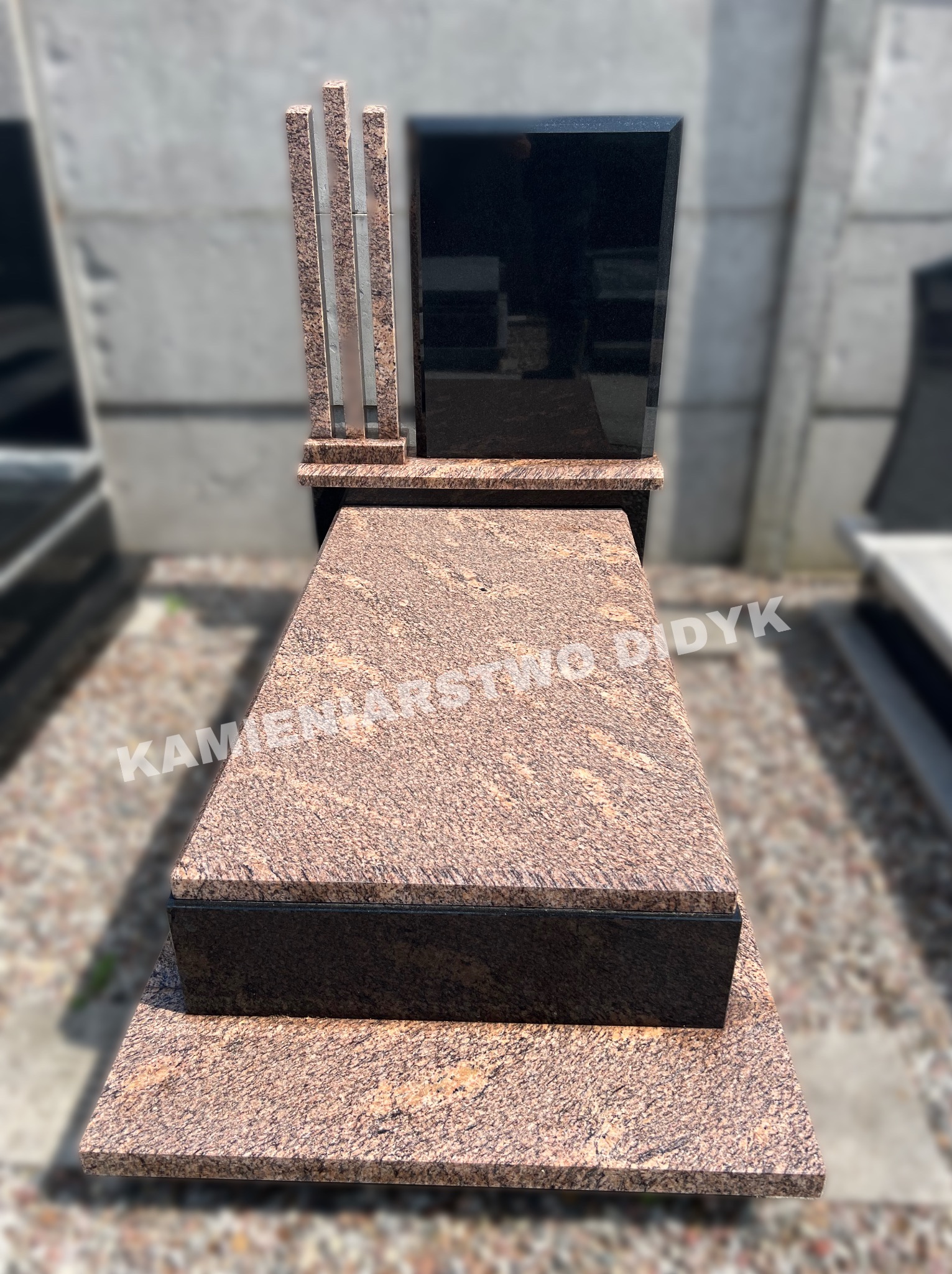 Granitowy pomnik z czarną tablicą i trzema kolumnami z jasnego granitu, widoczny na tle innych nagrobków.