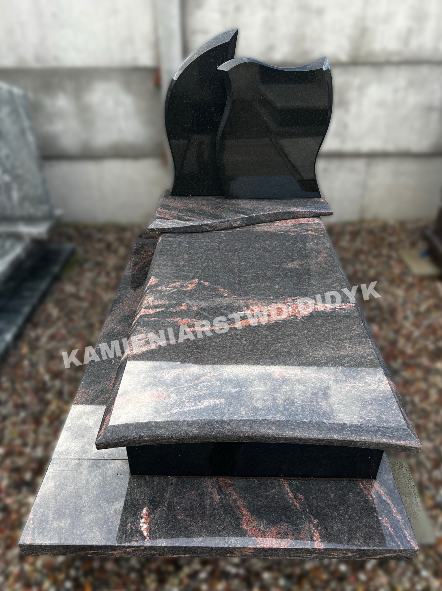 Elegancki, czarno-czerwony pomnik z ciemnego granitu, z wyprofilowaną górną płytą, ustawiony na żwirowym podłożu.