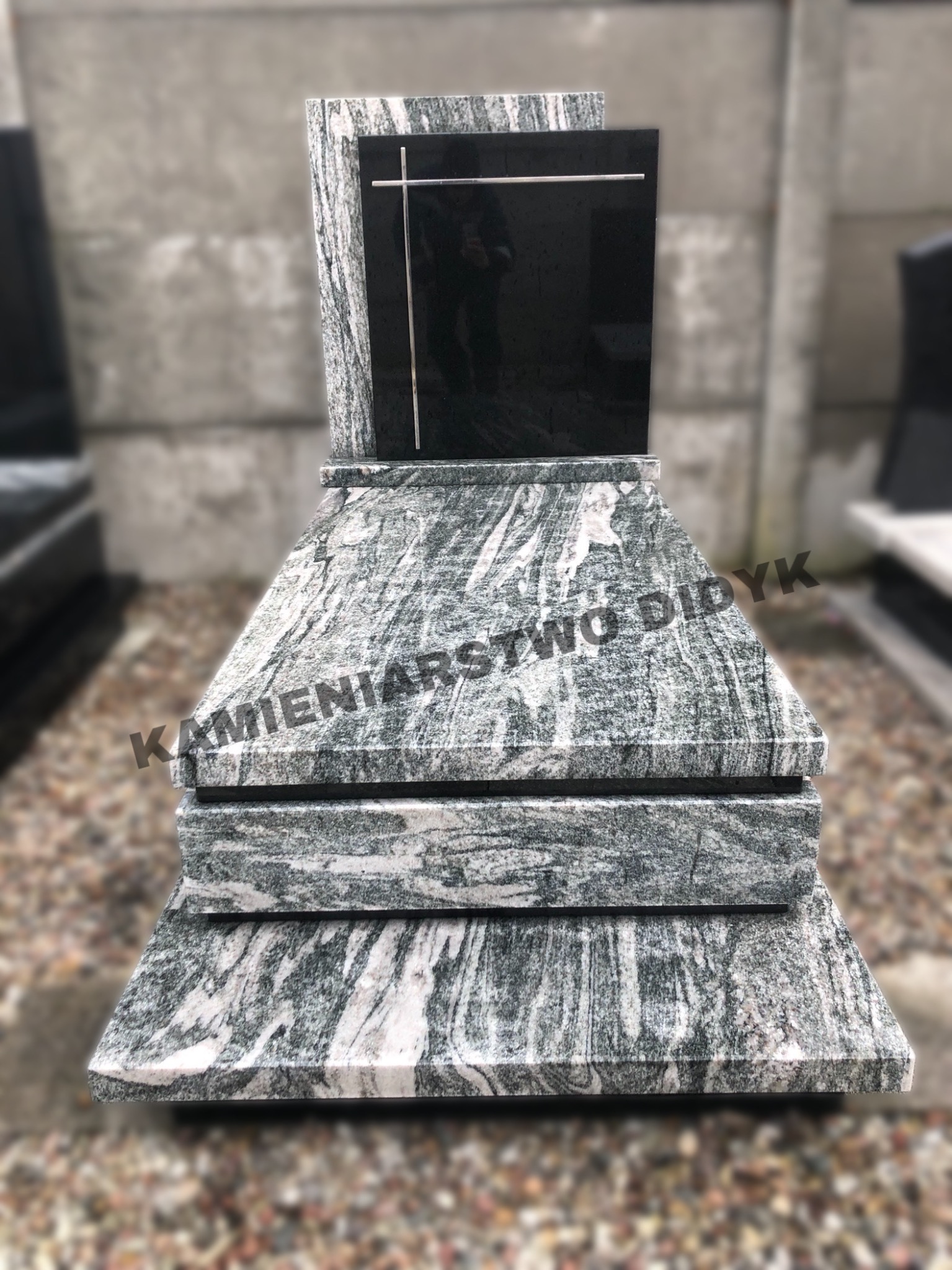 Granitowy pomnik z czarną tablicą i metalowym krzyżem, widoczne ziarna granitu w odcieniach szarości i bieli, na podstawie z jasnego kamienia.