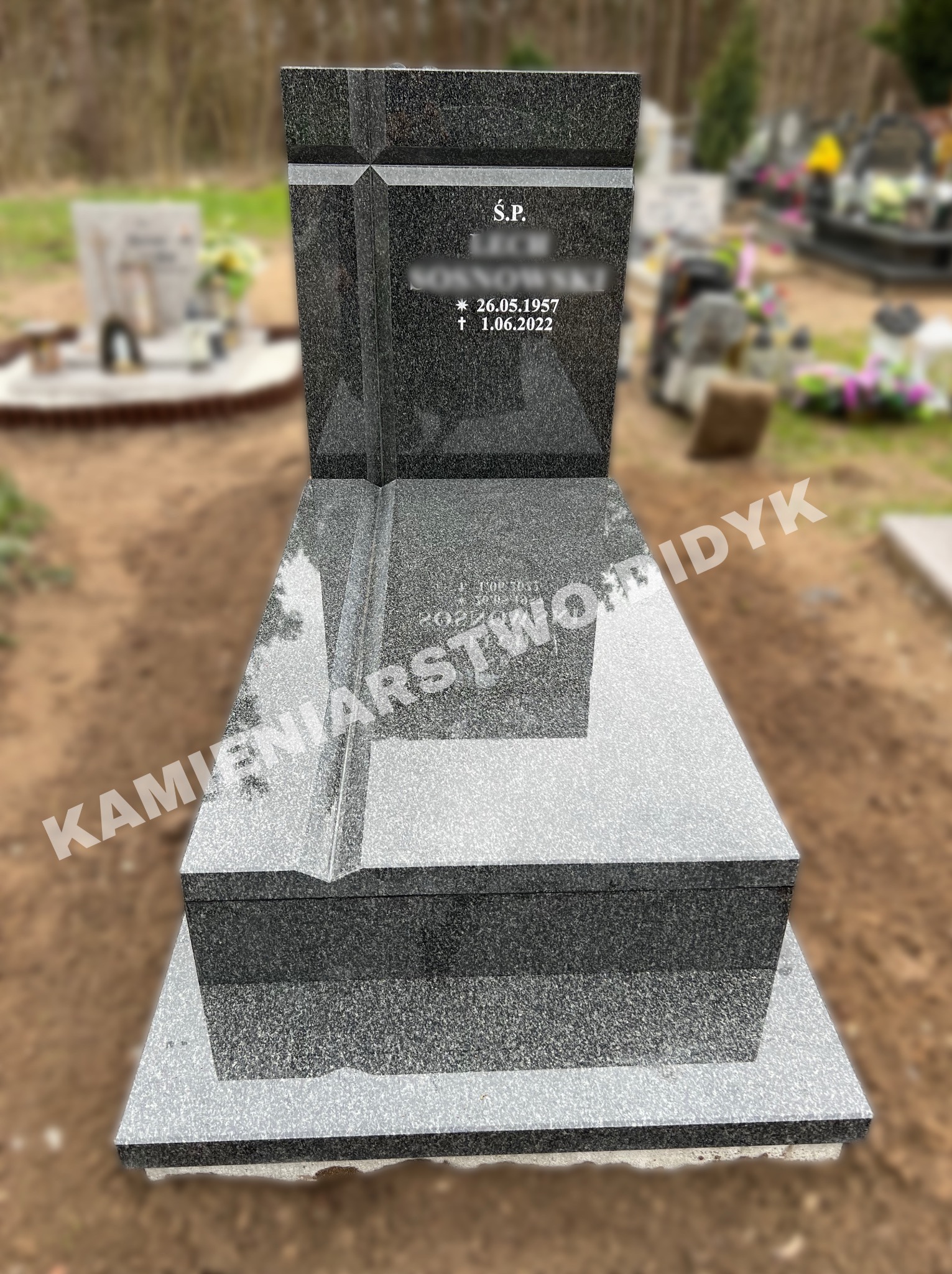 Granitowy pomnik z inskrypcją na cmentarzu, widoczne inne nagrobki w tle, spokojna atmosfera.