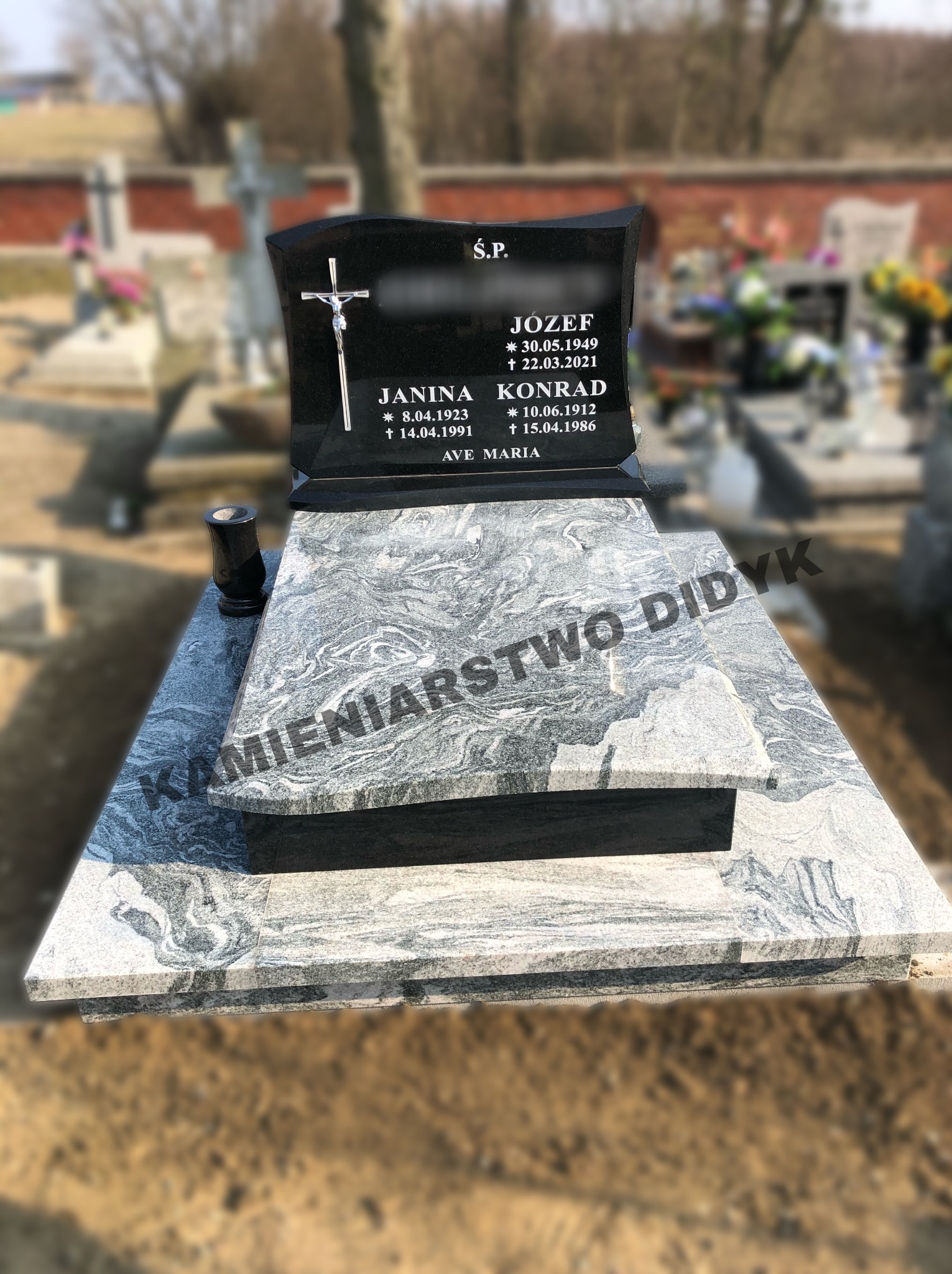 Czarny nagrobek z krzyżem i inskrypcjami na płycie z szarego, wzorzystego kamienia, na cmentarzu, z widocznym w tle innym nagrobkiem i drzewami.