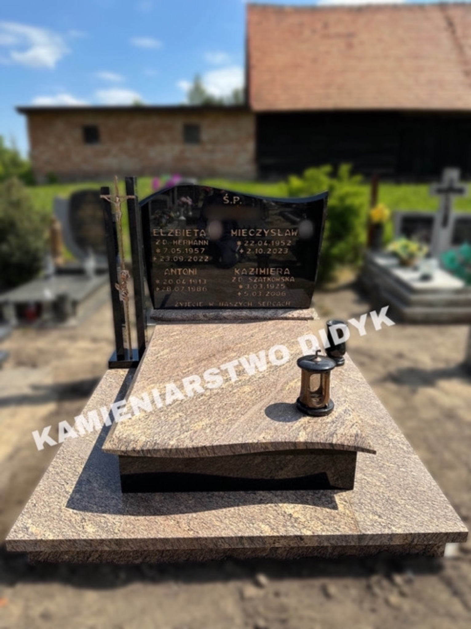 Granitowy pomnik nagrobny z czarną tablicą inskrypcyjną, ozdobiony metalowym krzyżem i lampionem, ustawiony na cmentarzu z rozmytym tłem drzew i budynków.