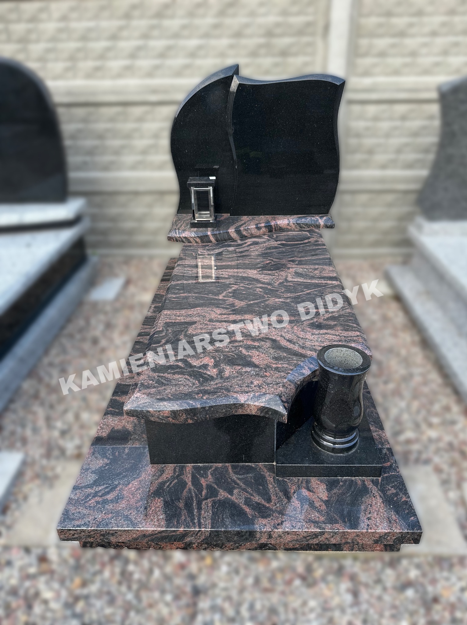 Czarny nagrobek z falistym wzorem, z wazą i lampionem, na tle jasnego muru